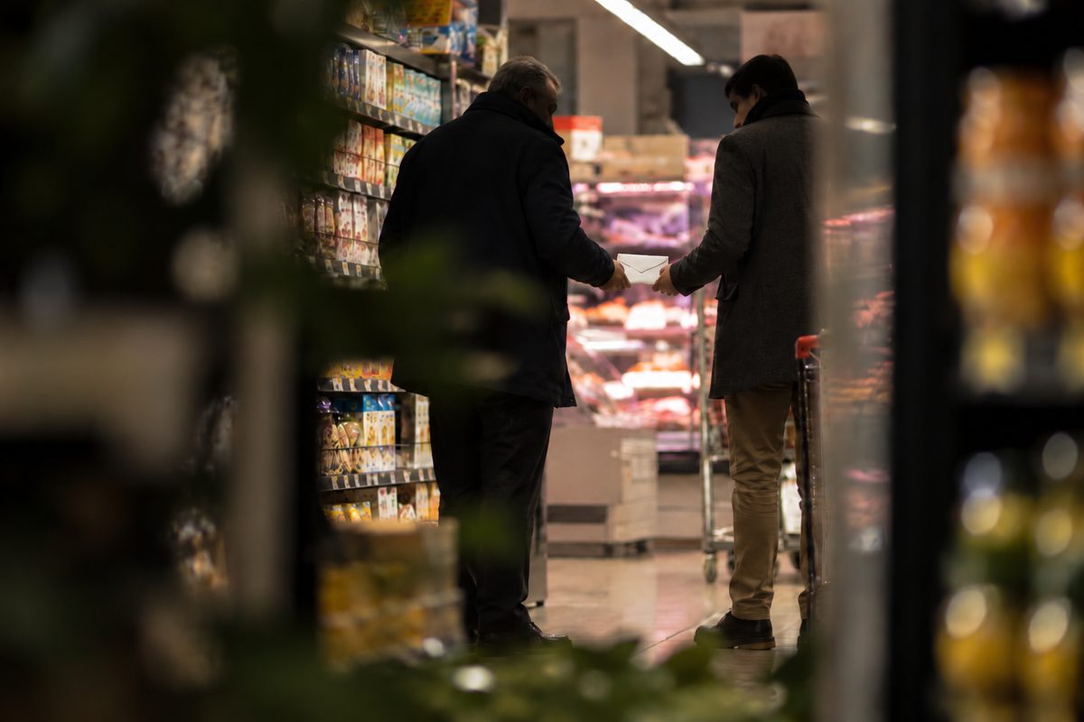 Due persone con i carrelli in una corsia di supermercato si passano con discrezione una busta